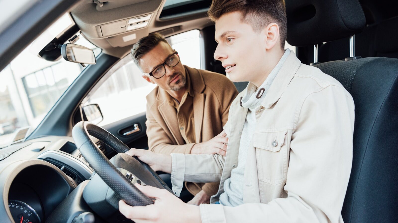A father coaching his teenage son at the wheel of a car.