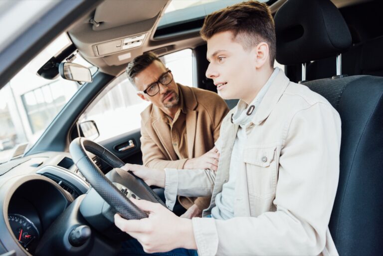 A father coaching his teenage son at the wheel of a car.